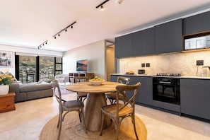 A modern kitchen with dark cabinets, a wooden dining table, and a large window overlooking a scenic view. - 270 Views, Modern, Bo-Kaap -Table Mountain Majesty Apt (Cape Town)