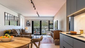A modern kitchen with a large island, a dining area with a wooden table and chairs, and a view of a cityscape through large windows. - 270 Views, Modern, Bo-Kaap -Table Mountain Majesty Apt (Cape Town)