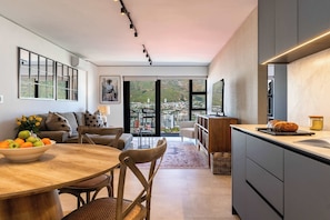 A modern kitchen with a large island, a dining area with a wooden table and chairs, and a view of a cityscape through large windows. - 270 Views, Modern, Bo-Kaap -Table Mountain Majesty Apt (Cape Town)