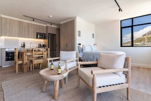 A modern kitchen with wooden cabinets, a dining area with a wooden table and chairs, a cozy seating area with a sofa and armchairs, and a large window with a view of a construction site. - Mountain Views, Shared Gym, CPT - York Urban Studio (Cape Town)