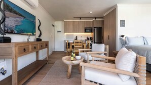 Una habitación de hotel moderna con una mesa consola de madera, una zona de estar con sofá y sillones, un área de cocina con microondas y refrigerador, y un televisor grande montado en la pared.