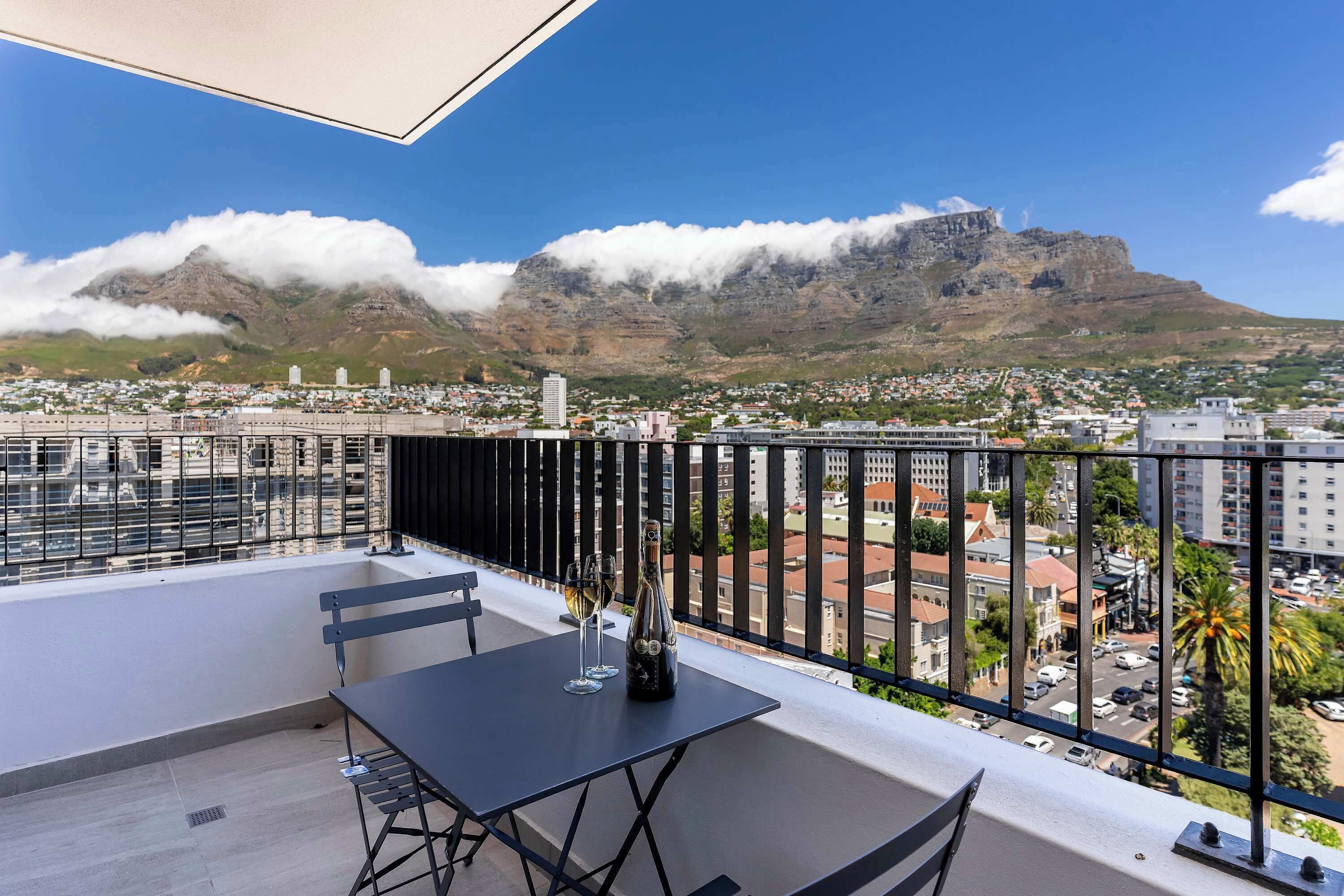 Un balcón con mesa y sillas, con vistas a un paisaje urbano que incluye montañas y nubes.