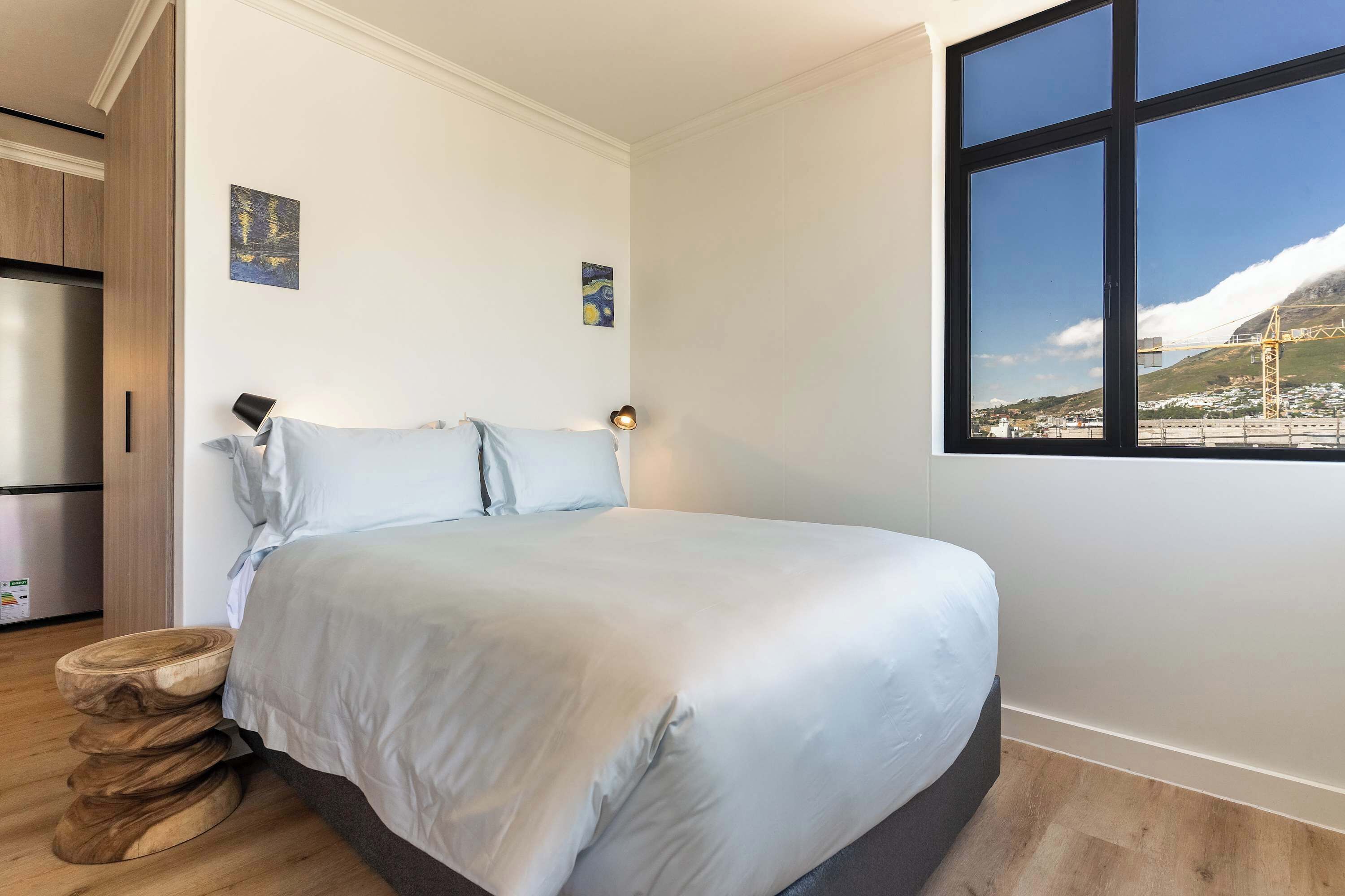 A bedroom with a bed, a wooden side table, a window with a view of a bridge and mountains, and a refrigerator in the corner.