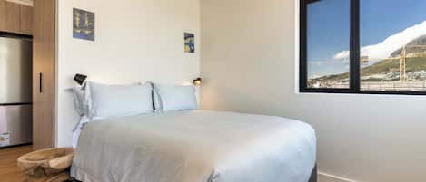 Un dormitorio con cama, una mesita de noche de madera, una ventana con vista a un puente y montañas, y un refrigerador en una esquina.