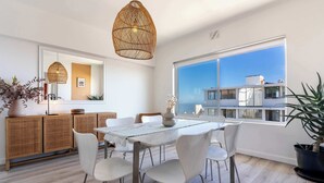 A modern dining area with a white table and chairs, a wicker ceiling light, and a large window with a view of buildings. - Perfect Location, Views - Bantry Bay Pearl Apt (Cape Town)