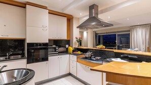 A modern kitchen with white cabinets, a stainless steel sink, a black countertop, and a large window with curtains. - Sunset Azure Apt - Panoramic Views, 2 min to beach (Cape Town)
