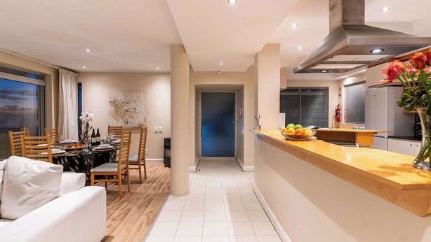 A modern kitchen with a wooden countertop, a dining area with a table set for four, and a living room with a sofa. - Sunset Azure Apt - Panoramic Views, 2 min to beach (Cape Town)