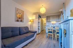 Ein modernes Wohnzimmer mit einer grauen Couch, einem Essbereich mit Holzsesseln, einer Küche mit weißen Schränken und einer Deckenleuchte.