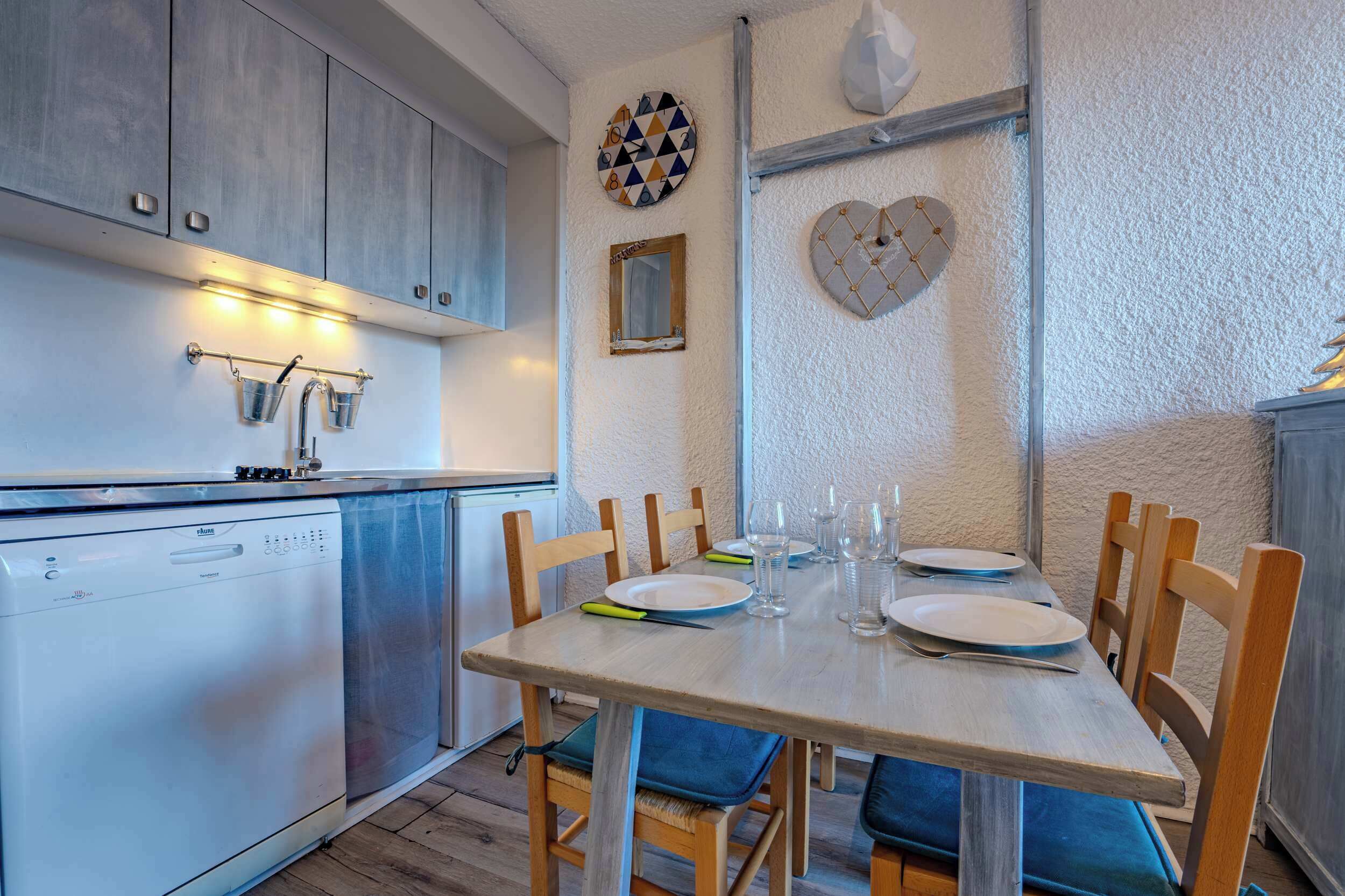 Uma cozinha compacta com lava-louças, cadeiras de madeira, uma mesa de jantar posta para quatro pessoas e arte decorativa na parede.