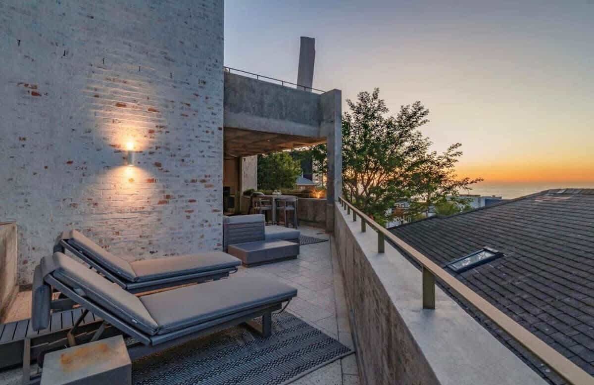 A rooftop terrace with lounge chairs, a dining area, and a view of the sunset over the ocean.