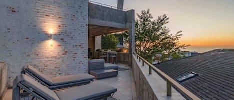 A rooftop terrace with lounge chairs, a dining area, and a view of the sunset over the ocean.