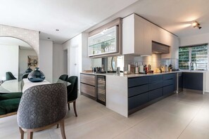 A modern kitchen with a central island, built-in appliances, and a dining area with a glass table and chairs. - Modern Apt, Views, Perfect Location - Bantry Alanda (Cape Town)