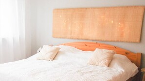 Ein ordentlich bezogenes Bett mit weißen Leinen und zwei Kissen, ein hölzernes Kopfstück und ein gewebter Wandbehang darüber.
