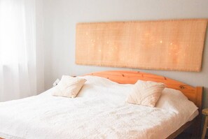 Ein ordentlich bezogenes Bett mit weißen Leinen und zwei Kissen, ein hölzernes Kopfstück und ein gewebter Wandbehang darüber.