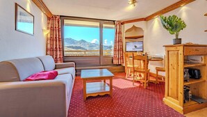 A room with a sofa, a small table, a wooden cabinet, a desk with a chair, and a large window with a mountain view. - Alpine Stay w/ Private Ski Views (Les Belleville)