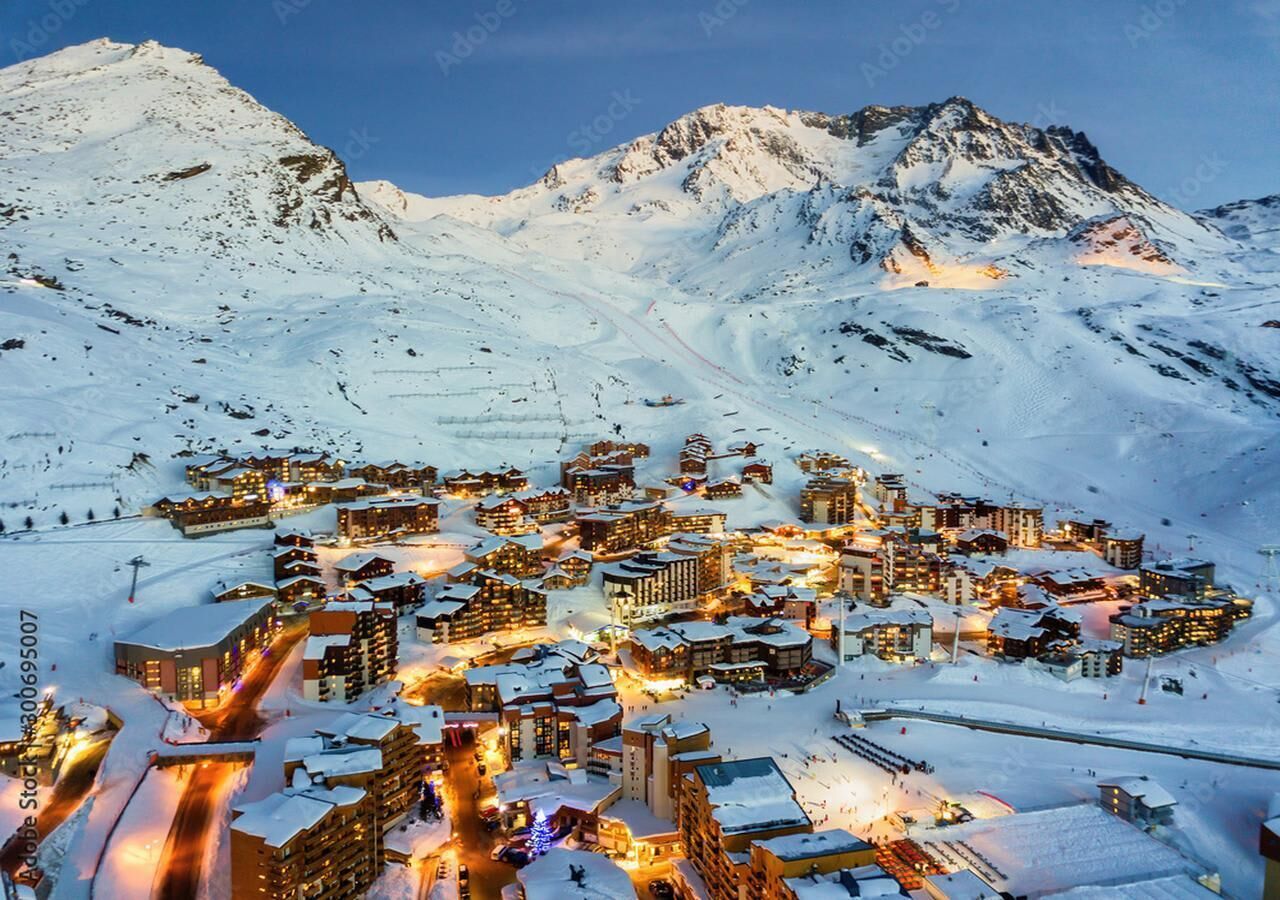 Un pueblo cubierto de nieve con edificios iluminados, probablemente un centro de esquí, que presenta un denso conjunto de cabañas y una vista despejada de las montañas circundantes.
