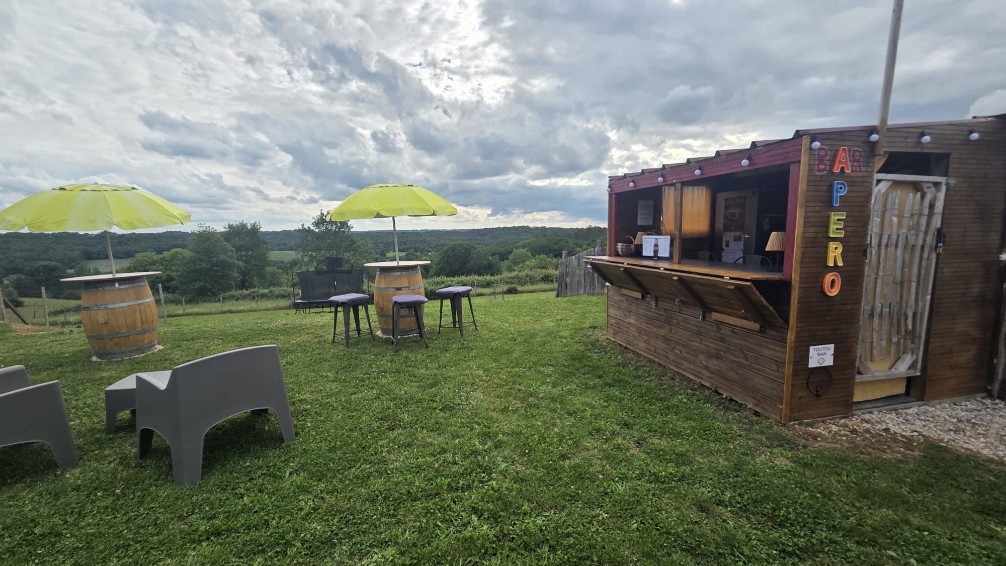 An outdoor seating area with tables and chairs, a wooden bar structure, and a view of the countryside.