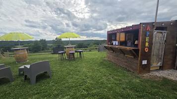 An outdoor seating area with tables and chairs, a wooden bar structure, and a view of the countryside.