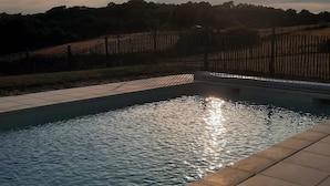 A swimming pool with a tiled deck, surrounded by a fence and trees, under a clear sky with the sun visible.
