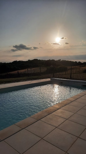 En swimmingpool med flisebelagte kanter, omgivet af et hegn og træer, under en klar himmel med synlig sol.