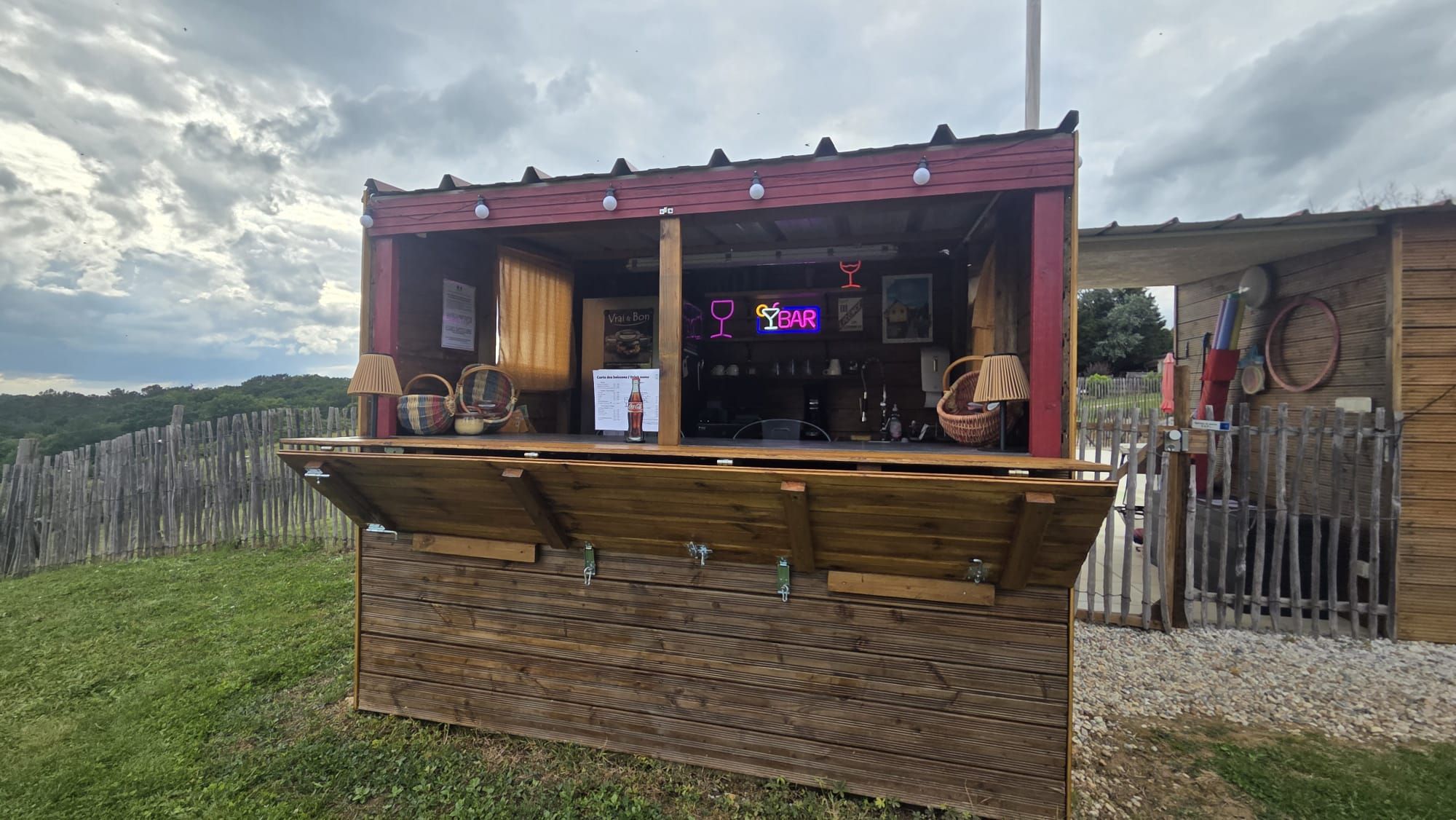 Een houten buitenbar met een rood dak, een neonbord en rieten manden.