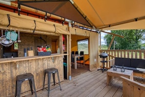 Un área de bar al aire libre con barra de madera, taburetes y una zona de estar con sofá y mesa.