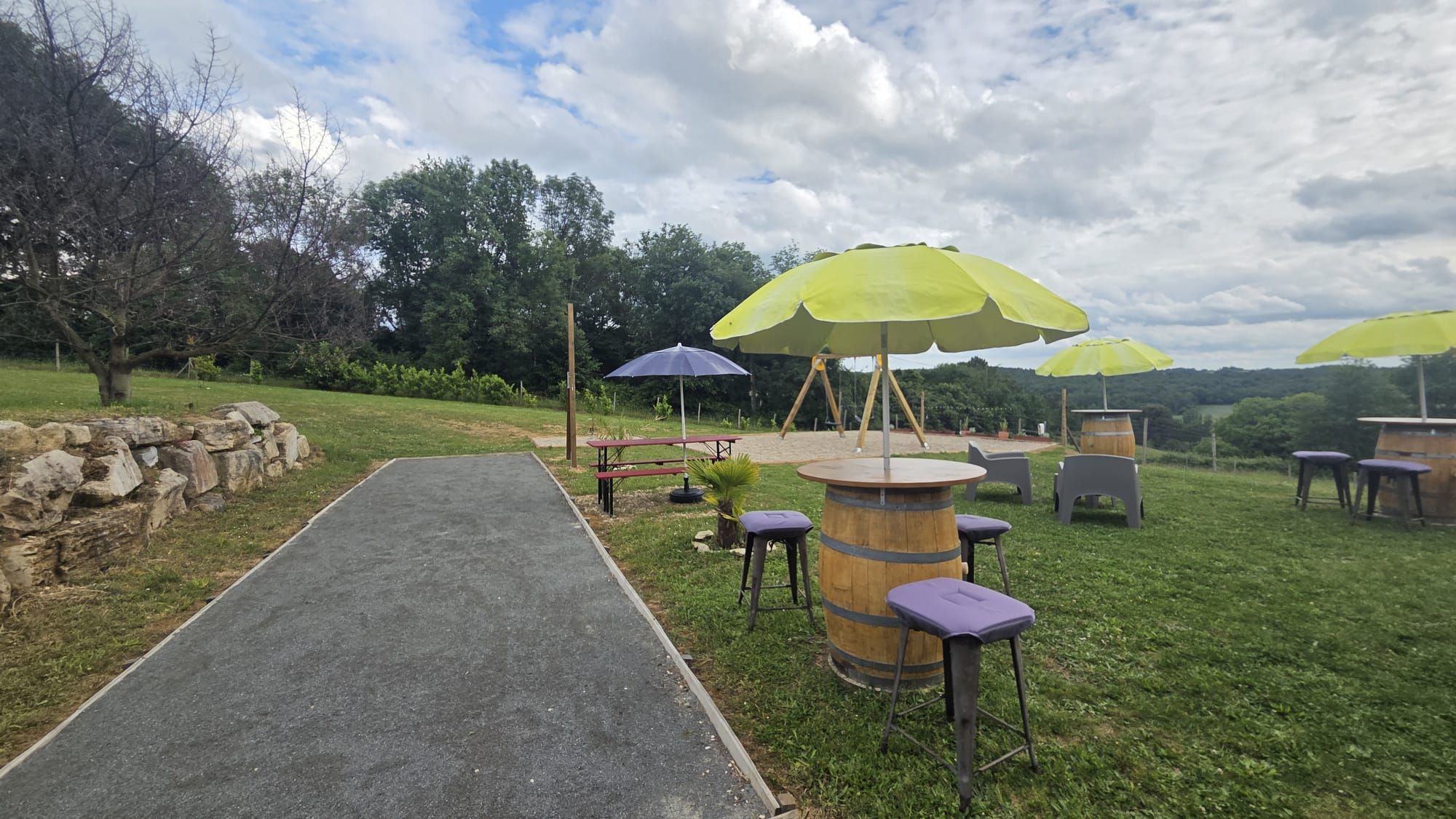 Un chemin pavé mène à un espace extérieur aménagé avec des tables et des tonneaux en bois, le tout entouré de verdure et d’arbres.