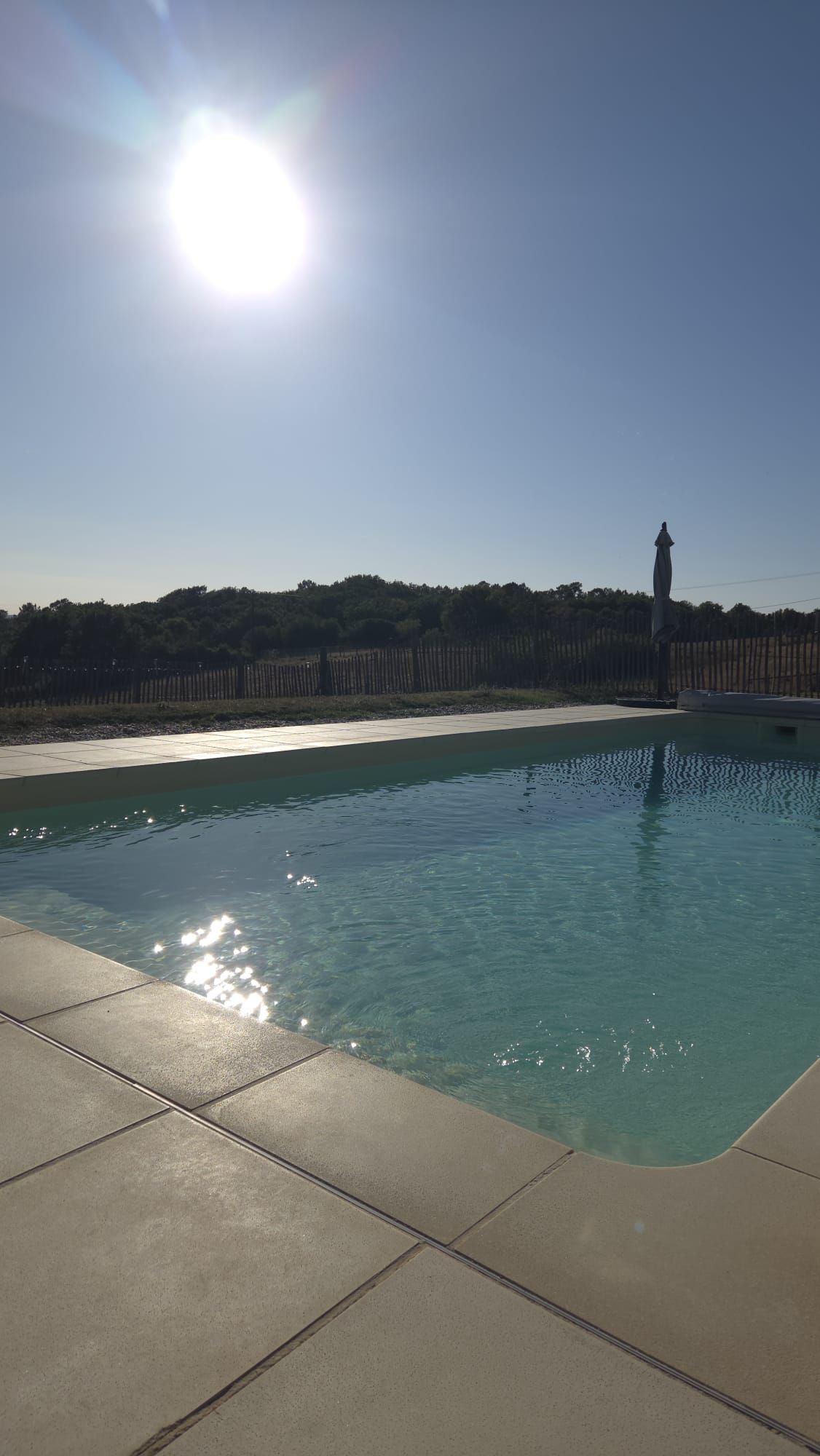 A swimming pool with clear water, surrounded by a tiled deck and a wooden fence.
