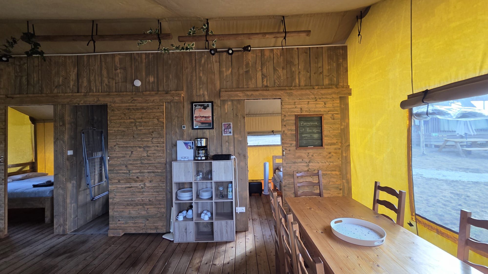 A rustic interior with wooden walls, a dining area with a wooden table and chairs, and a shelf with kitchenware.