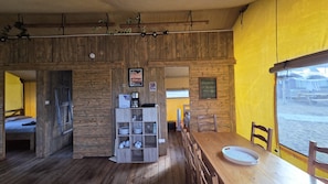 A rustic interior with wooden walls, a dining area with a wooden table and chairs, and a shelf with kitchenware.