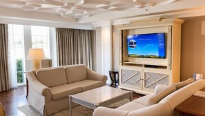 Una camera d'albergo con un divano color beige, un tavolino, una televisione su un mobile e una finestra con tende.