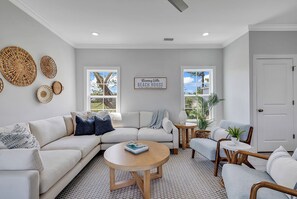 A modern living room with a sectional sofa, a round coffee table, and a wicker chair. There are two windows with views of trees and a door to the right. - Rosemary Villa, Heated Saltwater Pool, Volleyball Court, Golf Cart, Main ST (North Myrtle Beach)