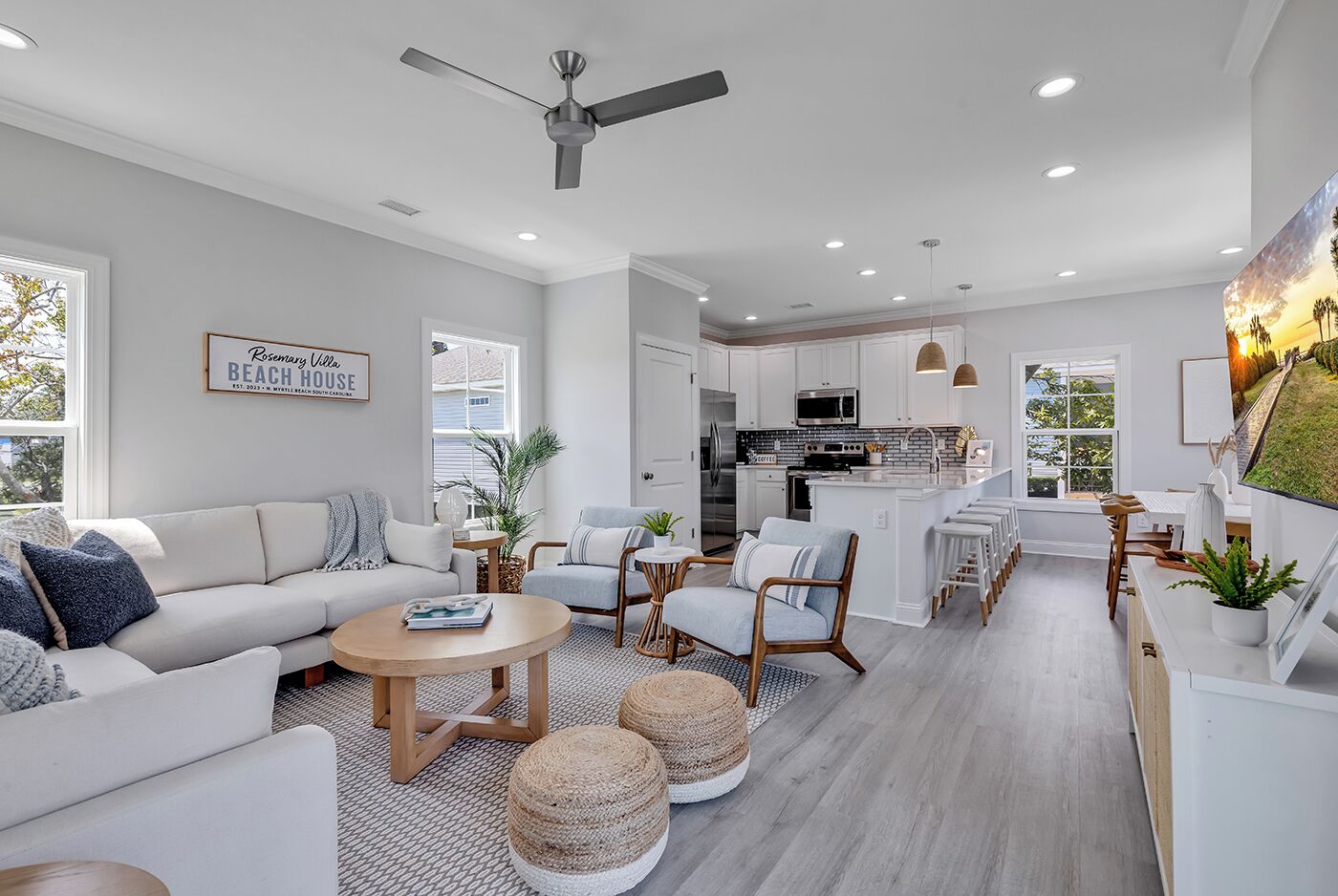 A modern living room with a sofa, coffee table, and chairs, a kitchen with white cabinets, and a dining area with stools.