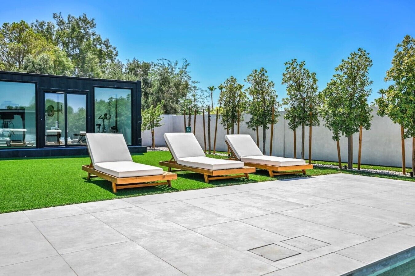 A modern outdoor area with three lounge chairs, a glass building, and a well-maintained lawn.