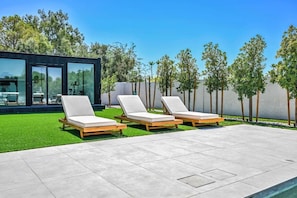 Een modern buitengebied met drie lounge stoelen, een glazen gebouw en een verzorgd gazon.