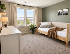 A bedroom with a bed, a bedside table with a lamp, a window with curtains, and a view of houses outside.