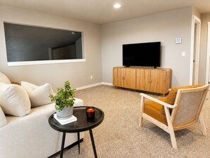 A living room with a sofa, a small table with a plant, a chair, a television on a wooden cabinet, and a large window.