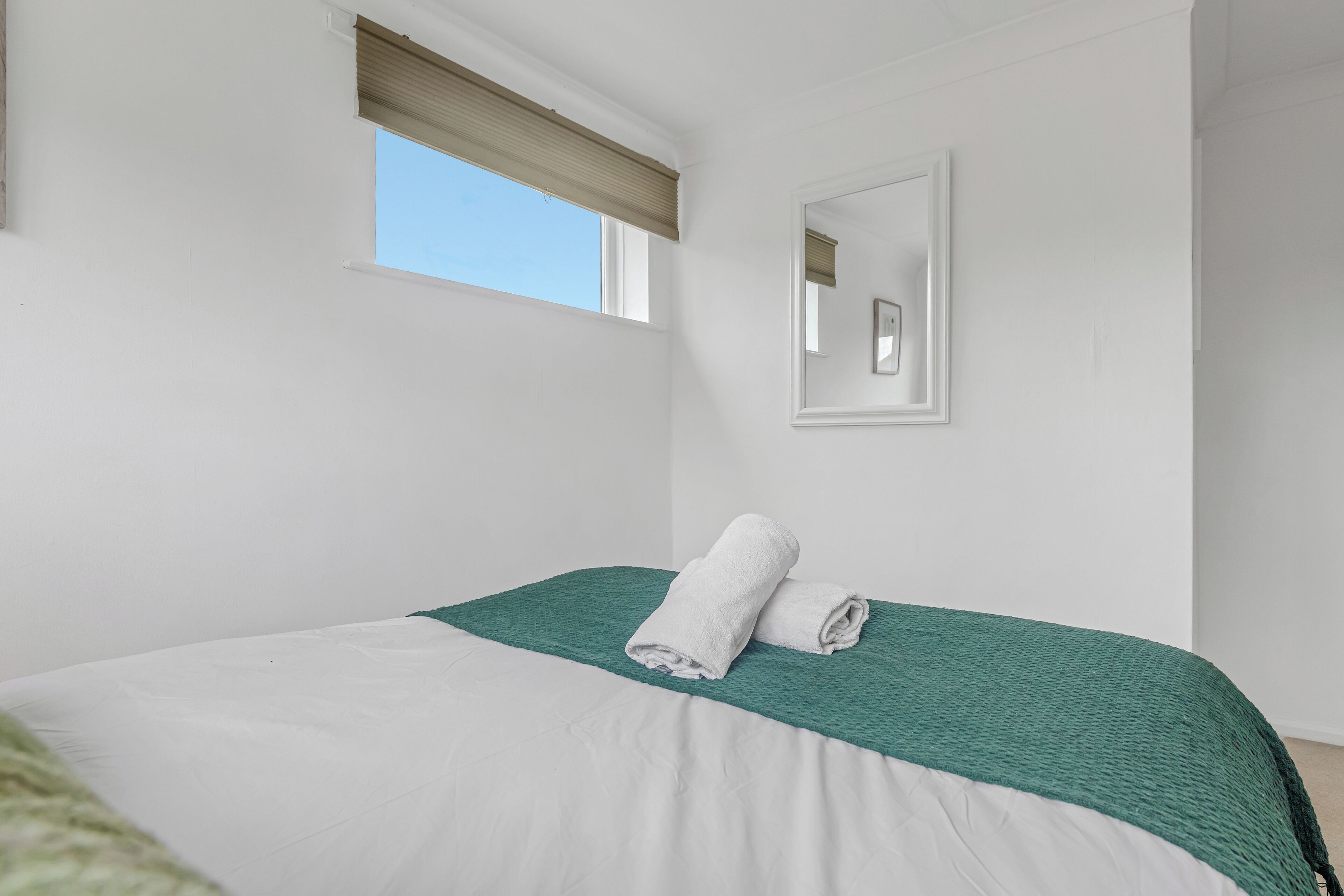 A bedroom with a bed, a window with a blind, a wall-mounted mirror, and a green textured throw blanket.