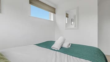 A bedroom with a bed, a window with a blind, a wall-mounted mirror, and a green textured throw blanket.
