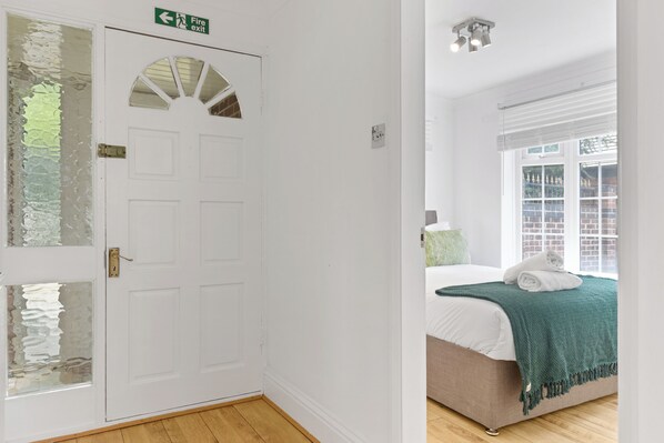 A room with a white door, a bed with a green blanket, and a window with a view of brick buildings.