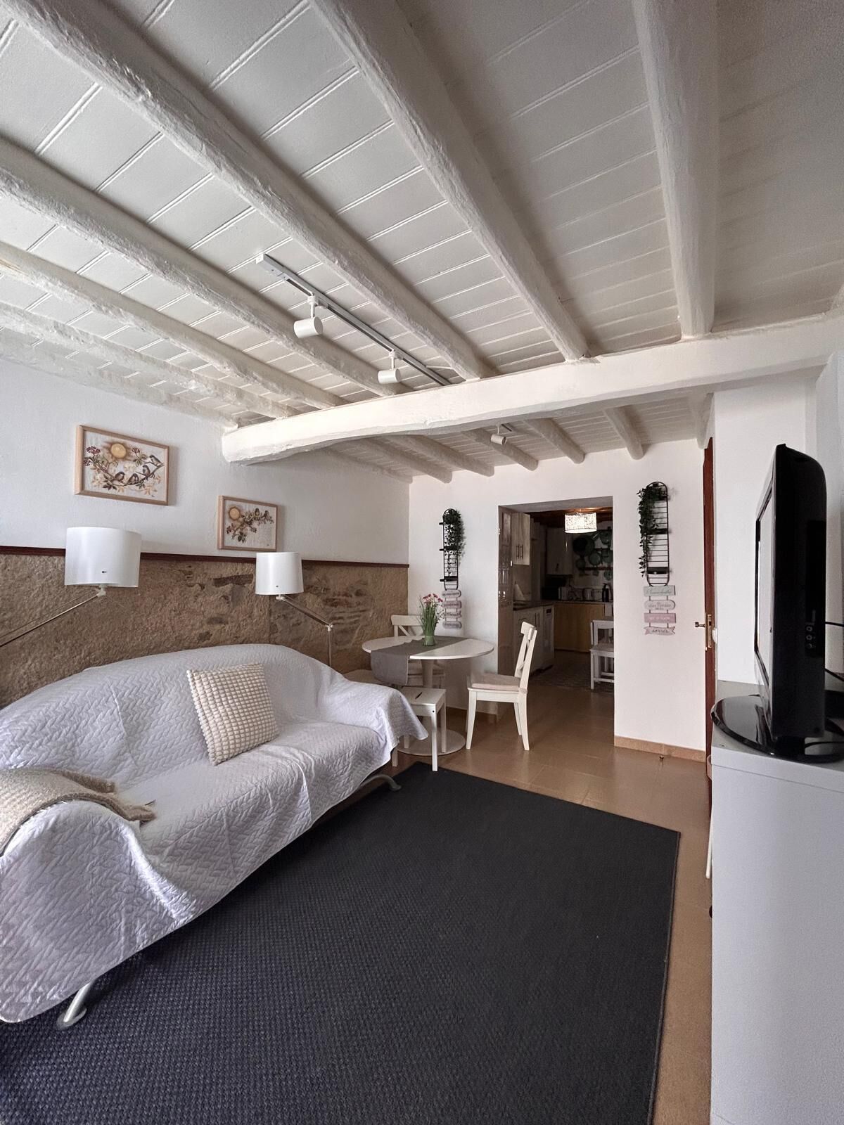 A bedroom with a bed, a desk, and a chair, a dining area, and a kitchen in the background.
