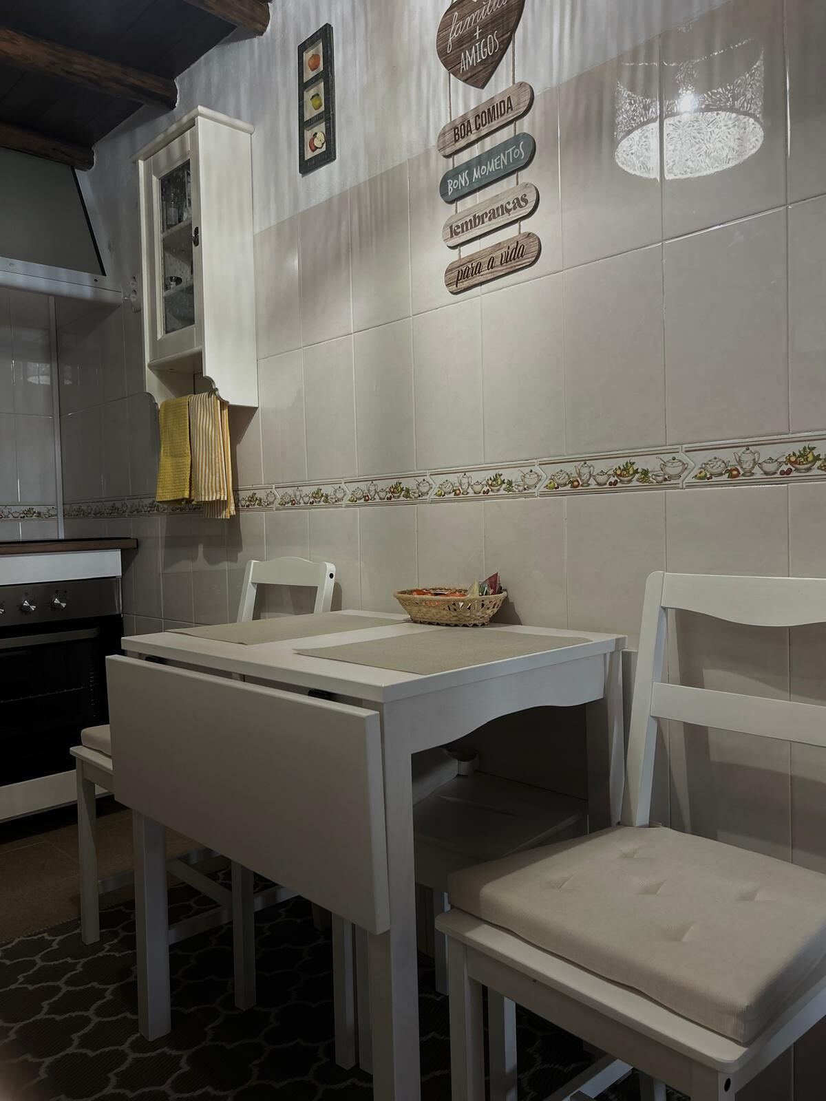 Una cocina con una mesa y sillas blancas, una estufa y un armario. Hay letreros en la pared con frases como 'família + amigos' y 'boda comida'.
