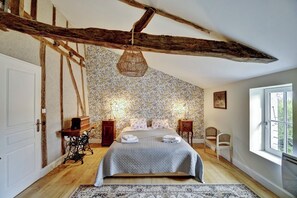 A bedroom with a bed, wooden beams, a patterned wallpaper, a hanging light fixture, and a window. - Vrbo Property (Plaisance)