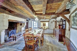 A rustic dining room with a wooden table set for four, surrounded by wooden chairs. A stone fireplace with a metal pot is visible on the left. - Vrbo Property (Plaisance)