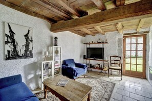 A rustic living room with exposed wooden beams, a television, a bookshelf, a blue sofa, and a patterned rug. - Authentic, renovated 3-bedroom house in the Périgord region (Plaisance)