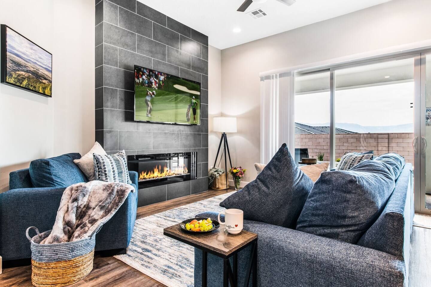 A modern living room with a fireplace, a flat-screen TV, and a large window offering a view of the outdoors.