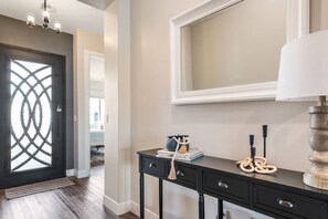 A hallway with a dark door, a light-colored wall, a mirror, a lamp, and a console table with books and decorative items.