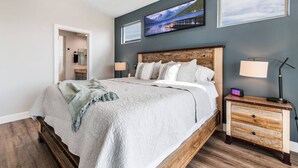 A bedroom with a large bed, a nightstand with a lamp, and a wall-mounted TV. - Grill and Chill by J and Amy BL 992912 (Mesquite)