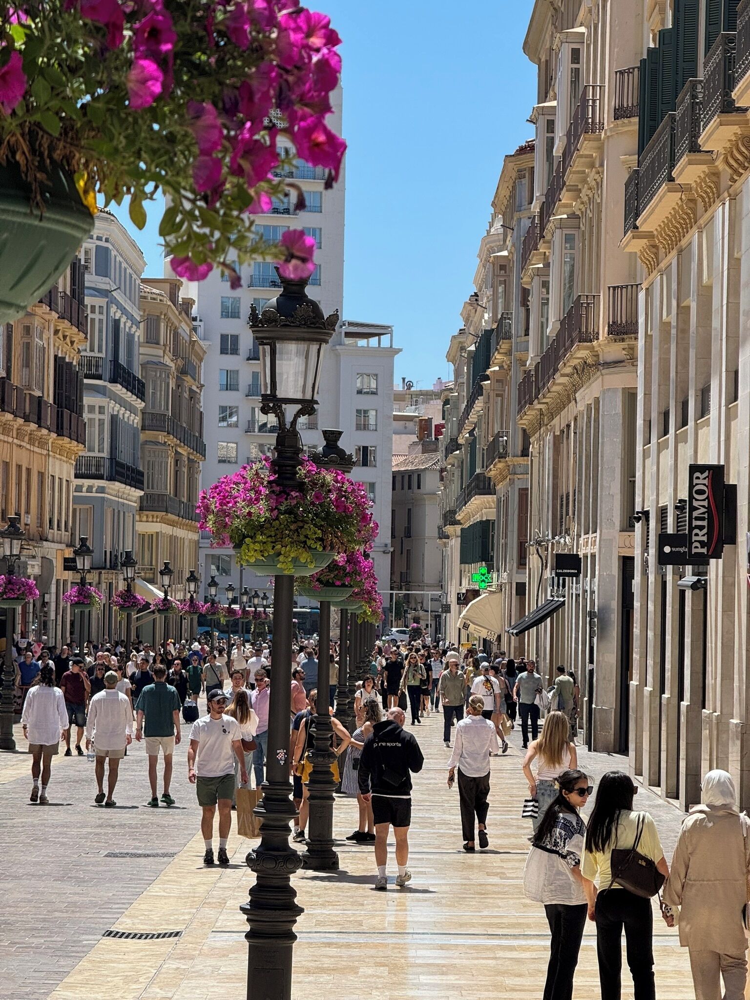 A bustling urban street with pedestrians, historic buildings, and a prominent street lamp with flower baskets.