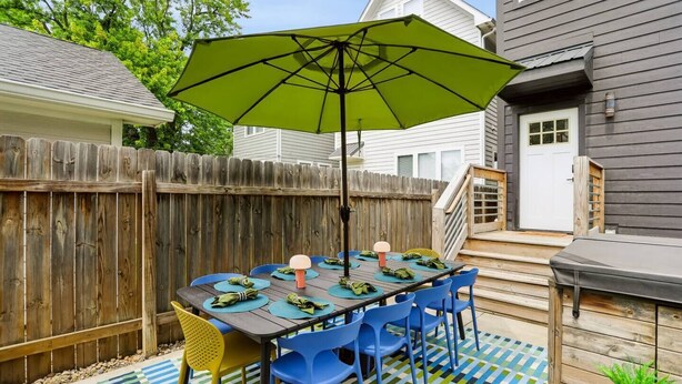 En udendørs terrasse med et bord dækket til fire personer, omgivet af blå stole, en grøn parasol og et træhegn.